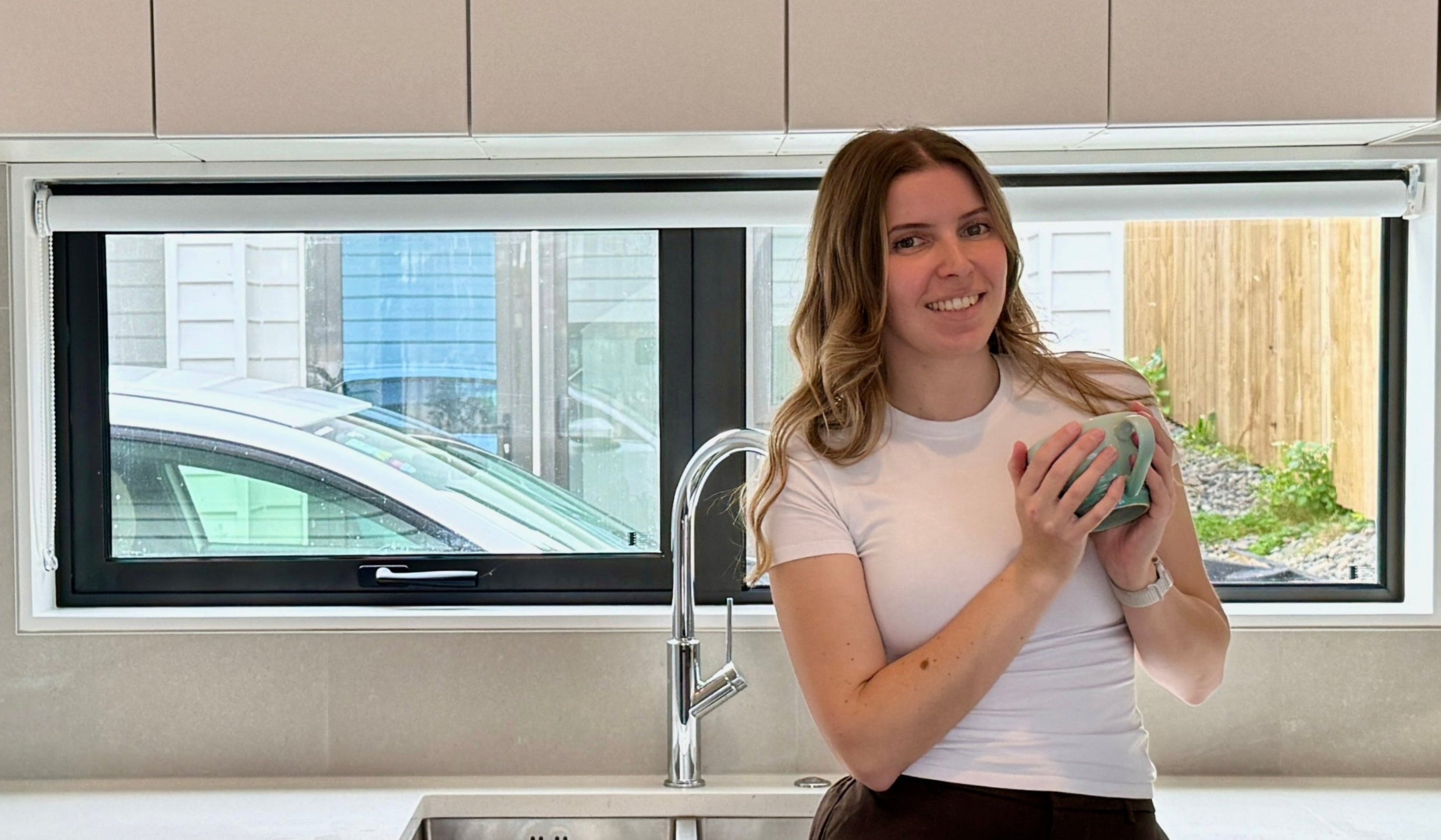 Woman holding a mug in a modern kitchen with a window showing a car outside.